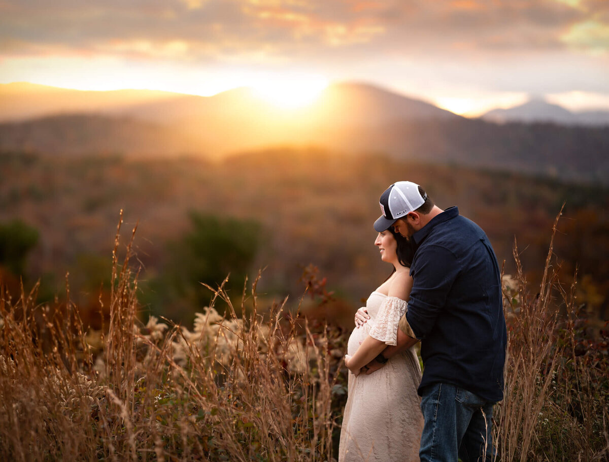 A couple stands together and embraces their baby bump as the sun sets behind the mountains