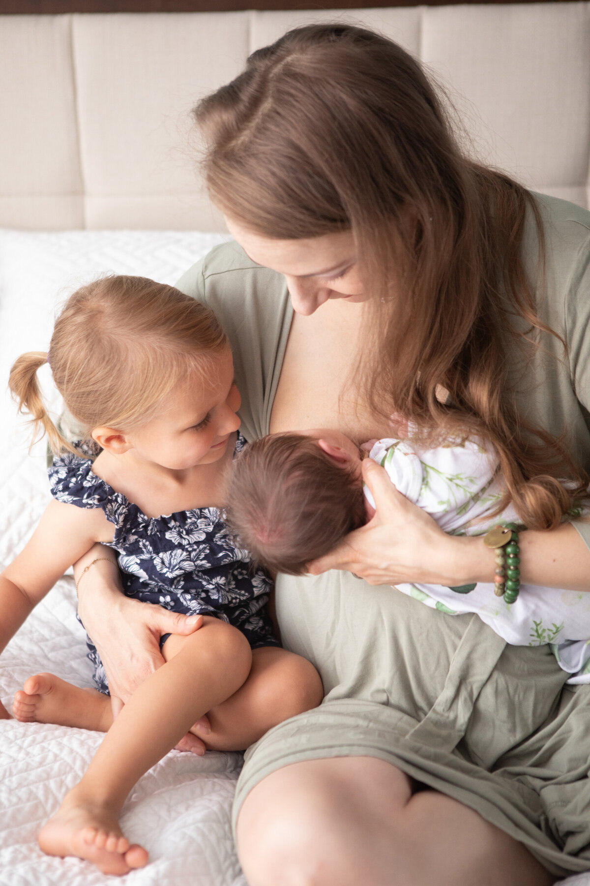 A woman in a light green dress nurses a newborn baby while her toddler daughter looks on newborn photographer White Plains Milestone Images