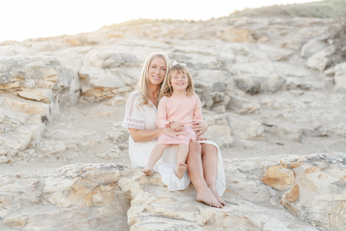 mom and daughter sitting on a rock at the beach and smiling at the camera
