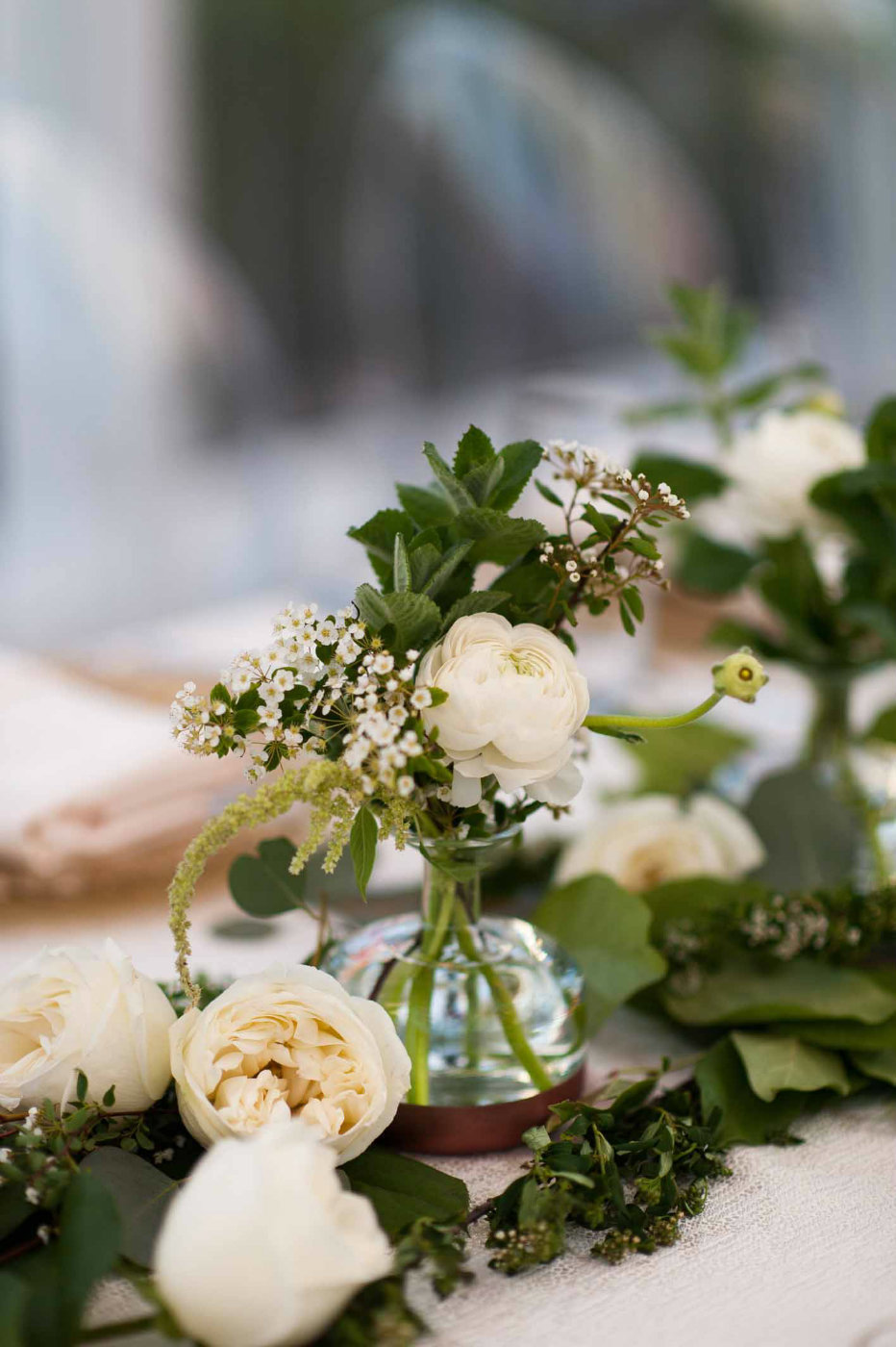 Small bud vases of greenery and white ranunculus are placed in the green garland on the head table.