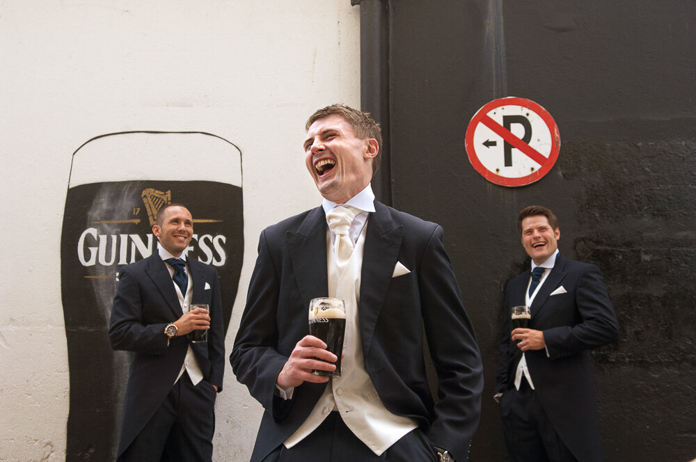 Groom and his groomsmen wearing black tailcoats and cream cravat laughing and holding pints of Guinness
