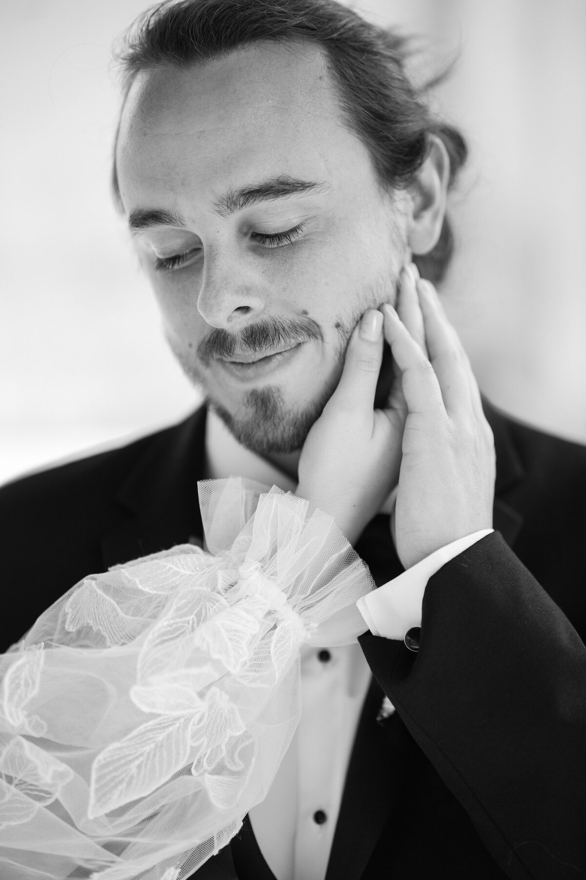 A black and white photo of a groom as his bride's hand caresses his face at their Charlotte, NC wedding.