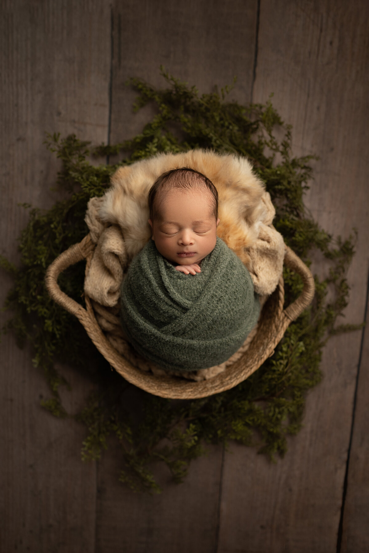 Katie Marshall, New Jersey's best newborn photographer captures a baby boy for his newborn fine art photoshoot. Baby boy is swaddled in an olive green knit with his fingers peeking out of the swaddle. He is sleeping peacefully and laying atop of faux furs in a basket.