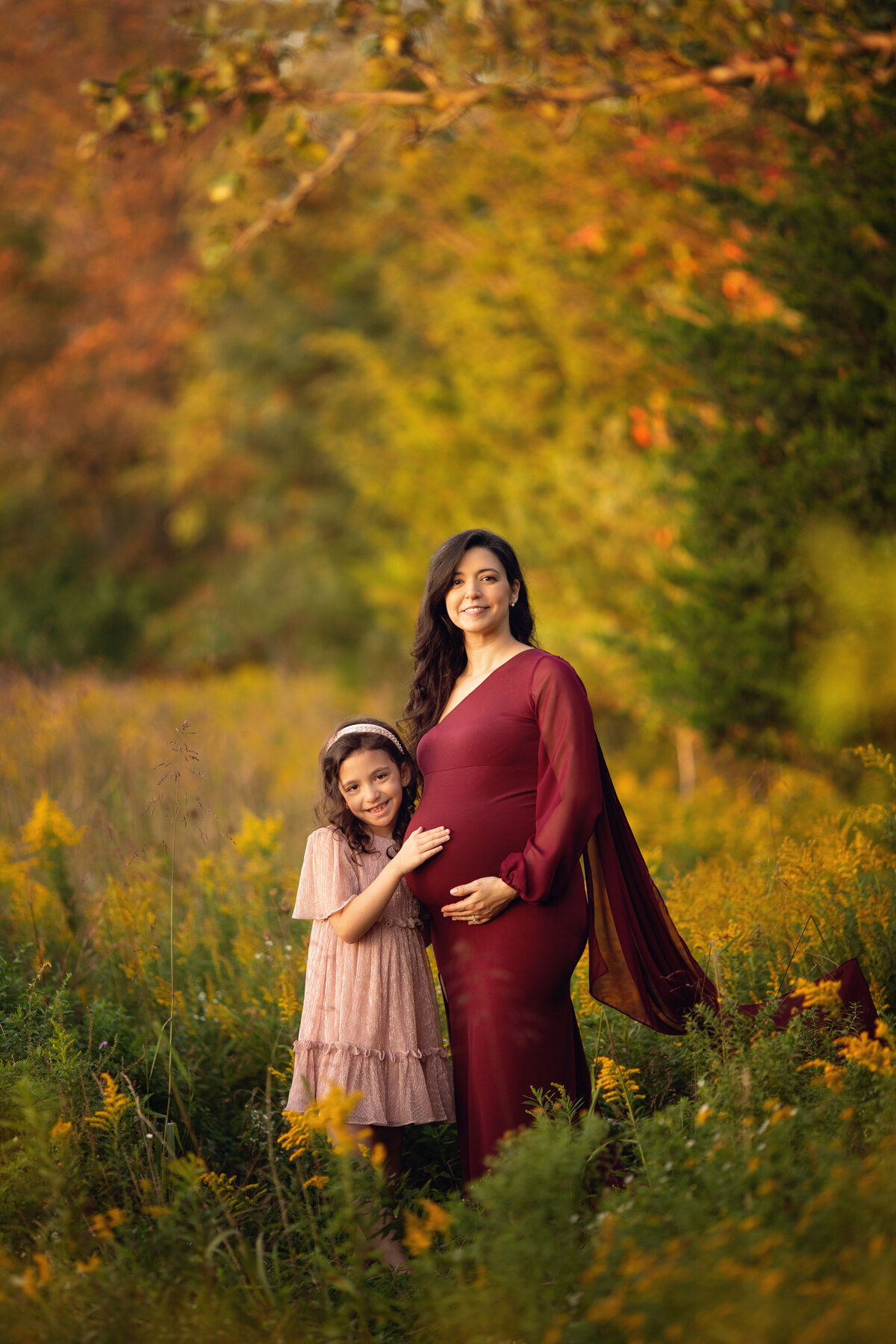 A pregnant mother in a red maternity gown stands in a field of tall grasses holding her bump with her toddler daughter in a pink dress