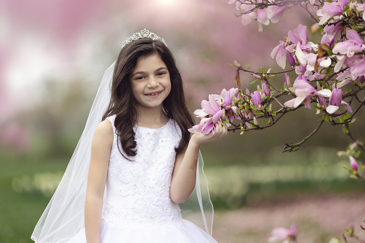 A smiling young girl stands with some pink flowers in a large white dress and tiara