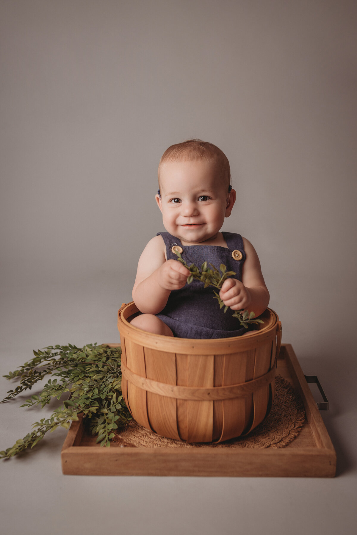 One year old baby boy sitting in a basket smiling, wearing a dark blue jumper on light gray backdrop