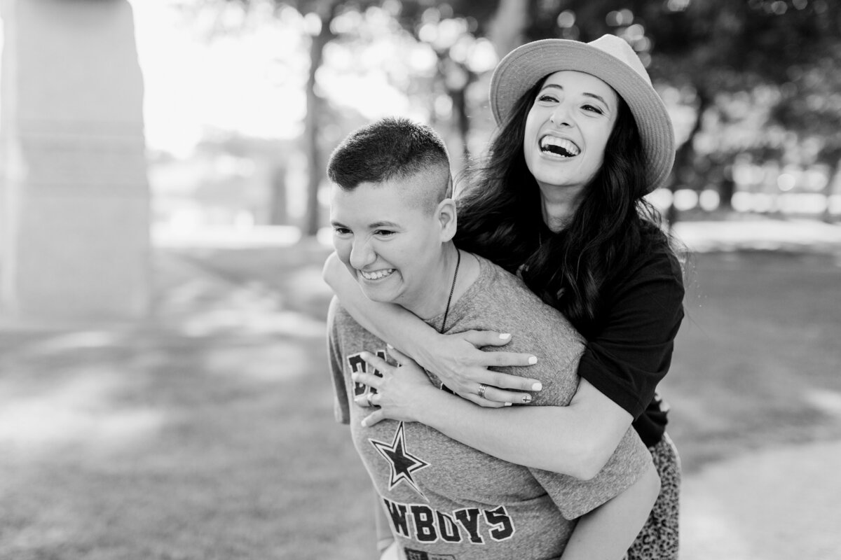 A black and white photo of a lesbian couple giving each other a piggyback ride during their engagement session in Arlington, Texas. The woman on the left is giving the piggyback ride and is wearing a Dallas Cowboys t-shirt. The woman on the right is receiving the piggyback ride and is wearing a dark top and a straw hat.