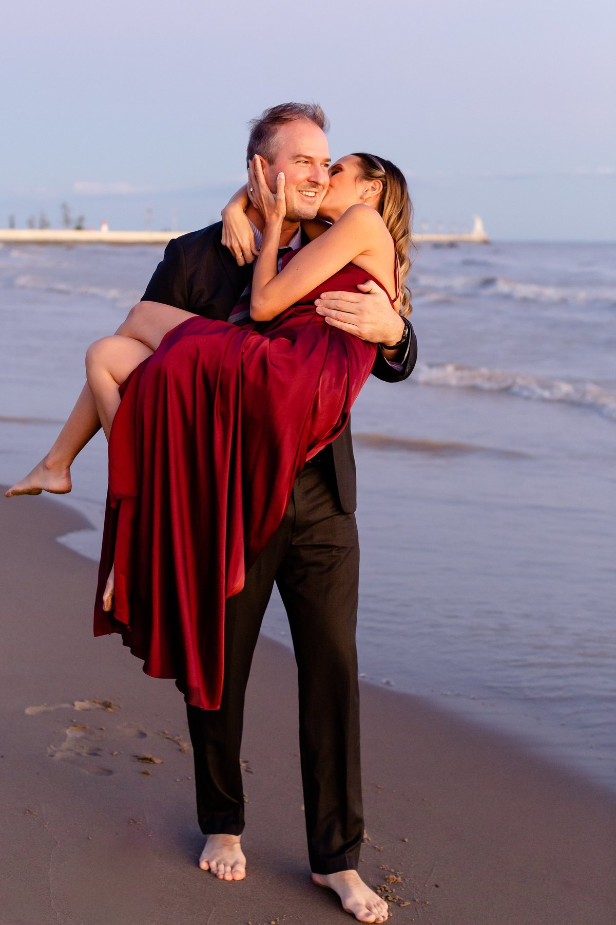Handsome-guy-carries-his-future-wife-wearing-a-fancy-red-dress-on-port-stanley-beach-during-their-sunset-engagement-session