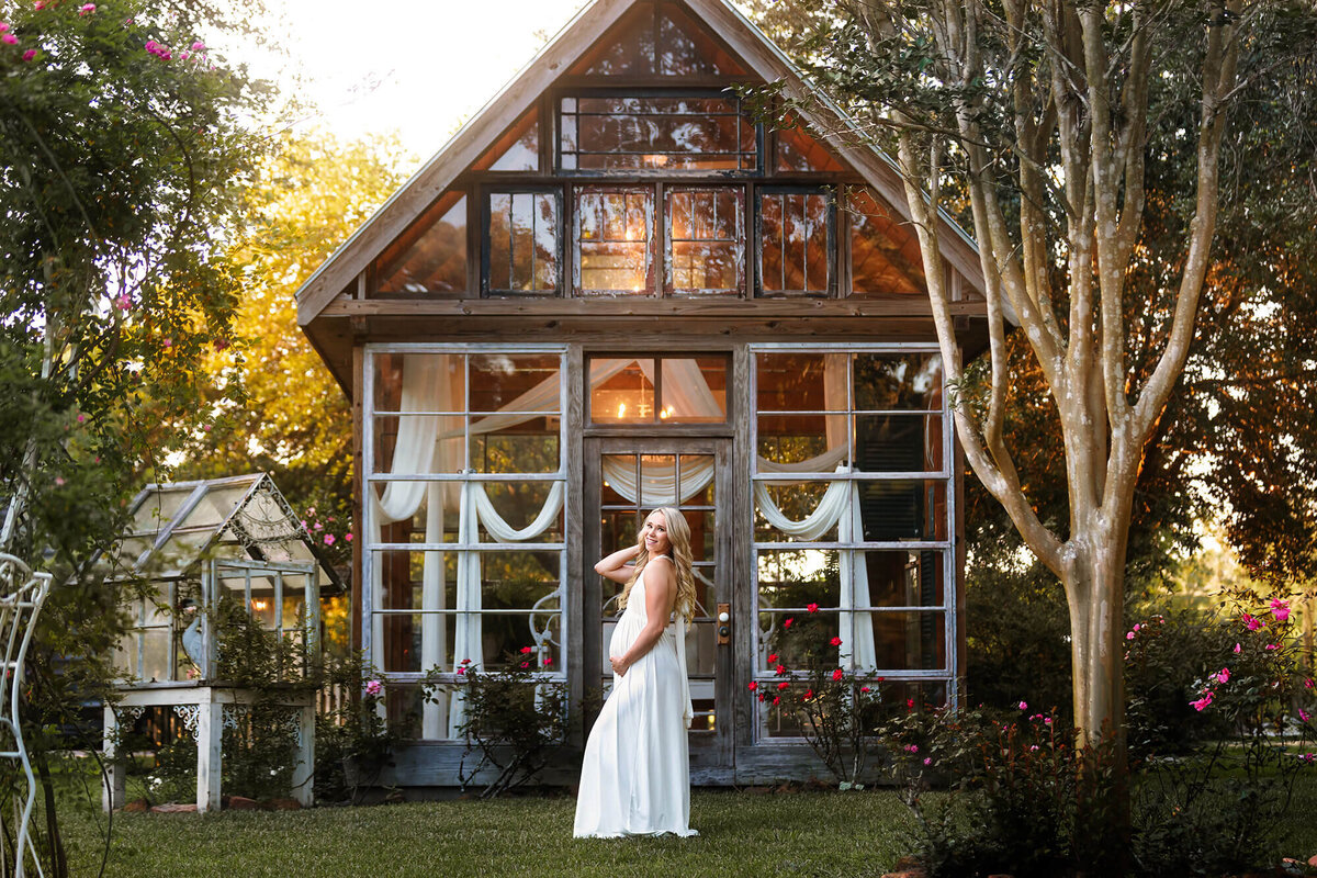 Stunning pregnant lady wearing a pure white dress in front of a greenhouse in the beautiful location in Angleton called Out Under the Trees.