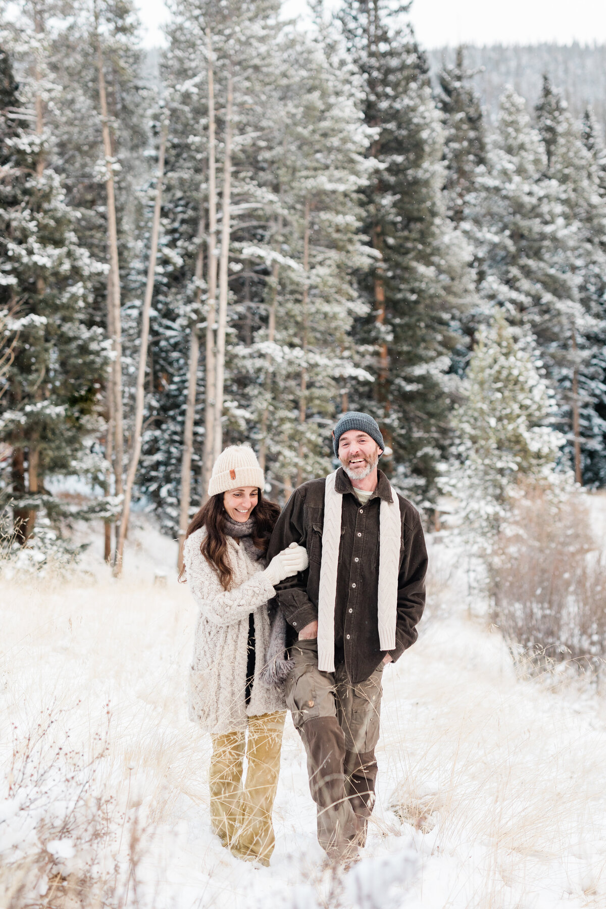 A husband and wife walk together in a winter wonderland of pine trees, brush, and lots of snow