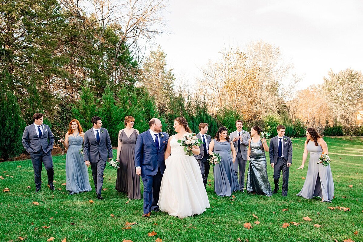 The bride, dressed in a long lave gown and veil and the groom dressed in a dark navy blue suit and tie stand in front of their bridal party dressed in a variety of varying shades of blues and grays as they stand outside at Sycamore Farms for an autumn wedding in tennessee