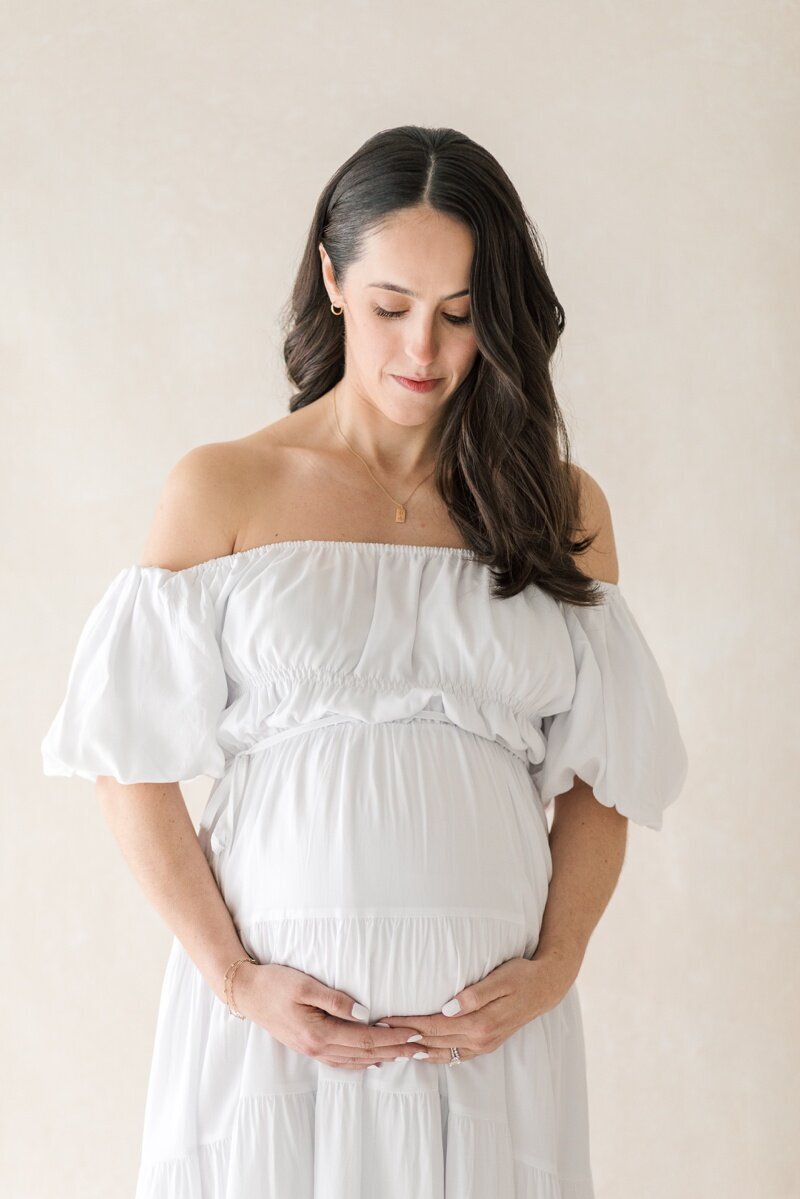 A pregnant woman wearing a white off the shoulder dress holds her stomach as she gazes down at it during her Lawrenceville NJ Maternity Photography session