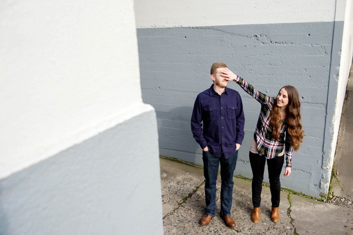 a fun and silly couple stands in front of a gray and white wall and cover each others eyes