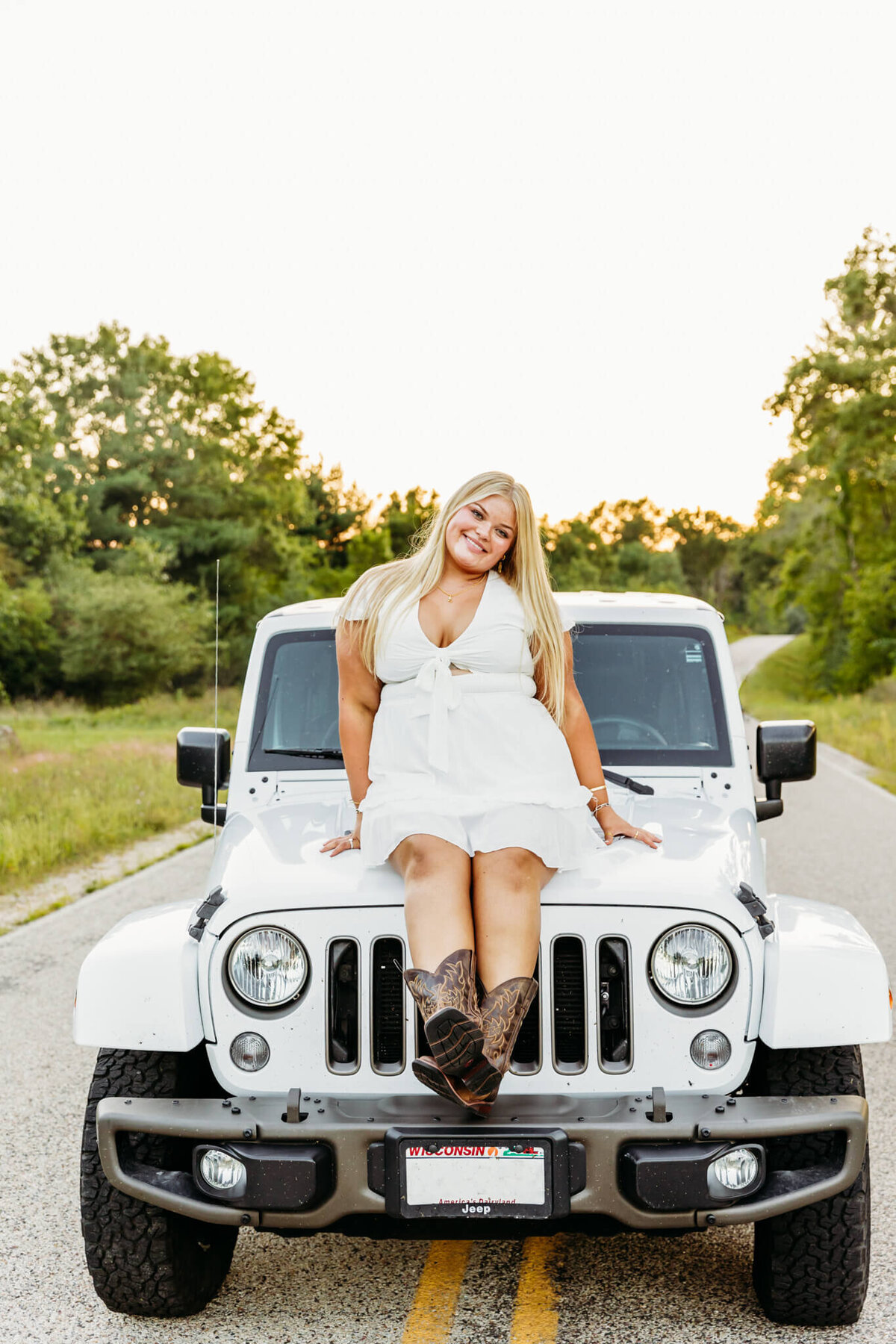 gorgeous high school senior girl in a white dress and cowboy boots sitting on the hood of her white jeep at sunset near Green Bay