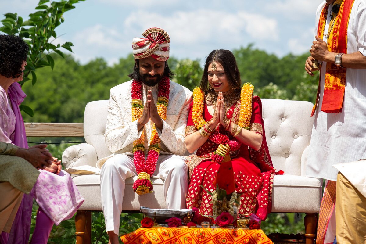 A bride and groom in traditional Indian attire sit side by side, performing rituals with joined hands at an outdoor wedding ceremony coordinated by a wedding planner in Des Moines.