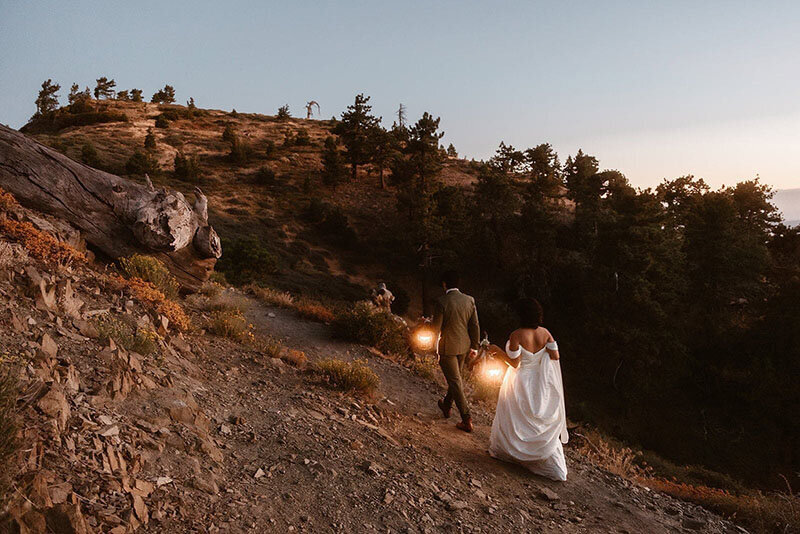 A bride and groom hike a winding mountain path at dusk, carrying lanterns as the light fades.