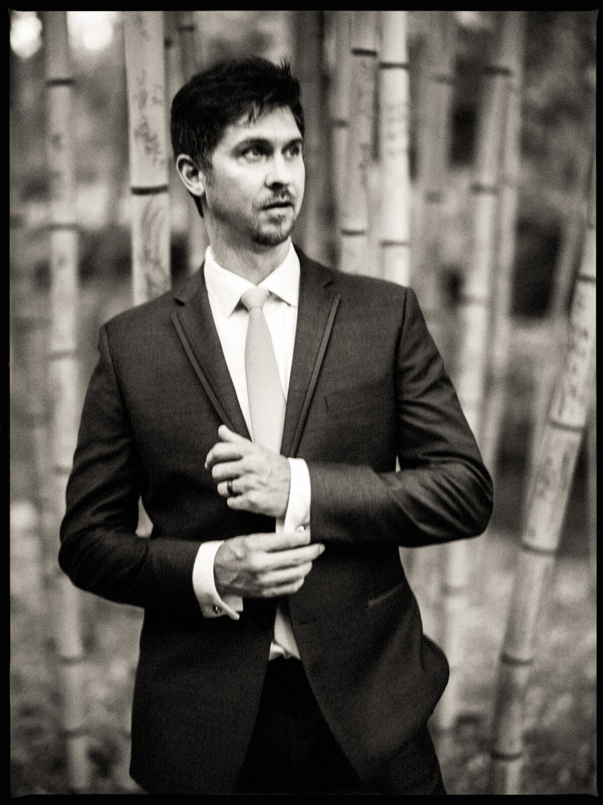 A groom in a dark suit adjusting his cufflink, looking thoughtful amidst a backdrop of soft-focus bamboo