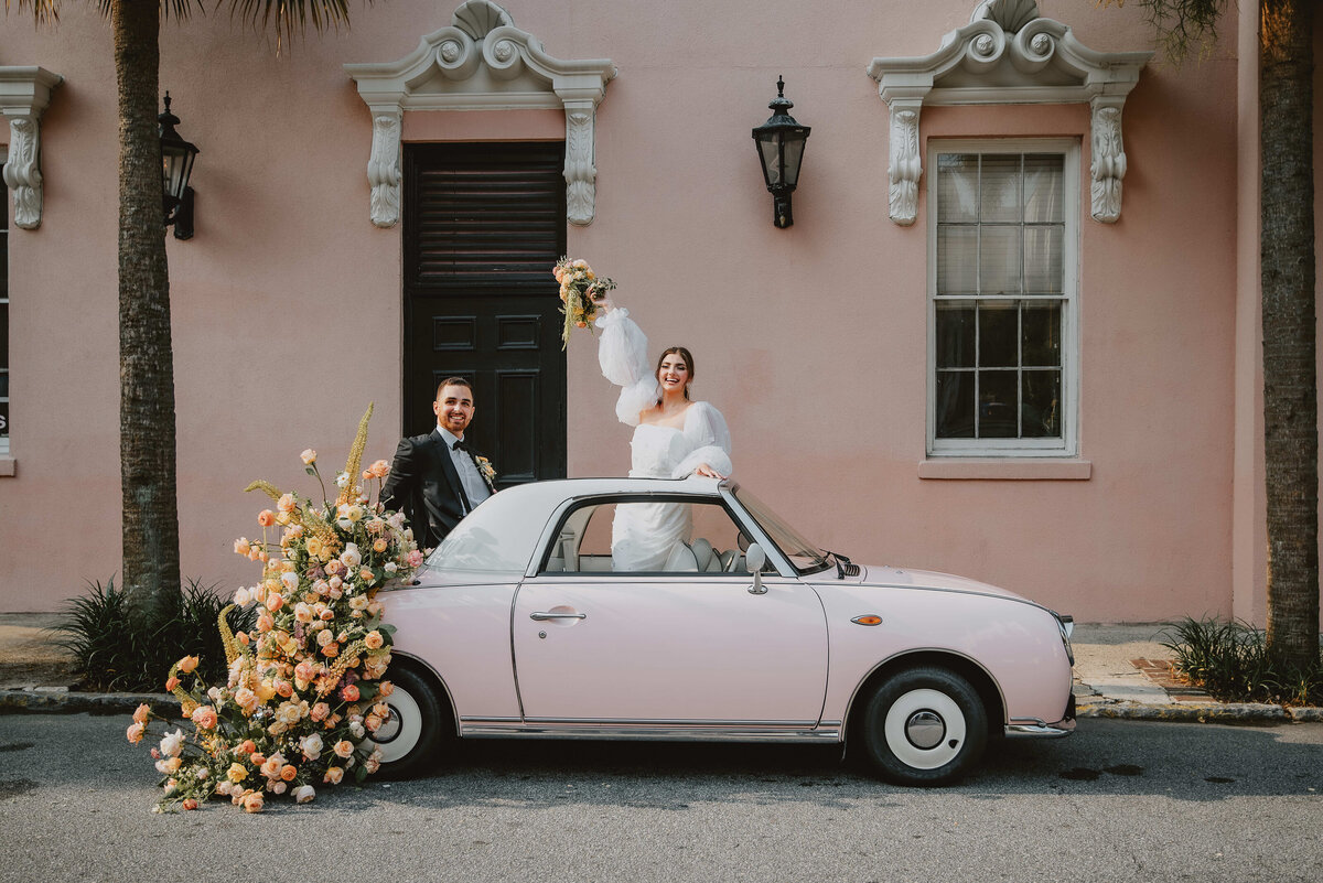 A couple in wedding attire stands with a vintage pink car adorned with flowers. The joyful scene is set against a pastel building, evoking romance and celebration.
