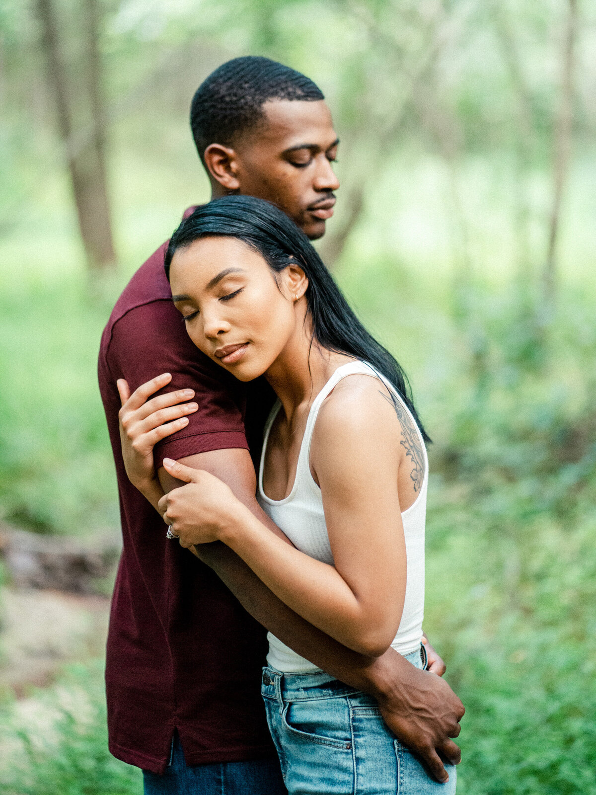A man and woman embracing in a forest, with the man's eyes closed and the woman's head resting on his shoulder.