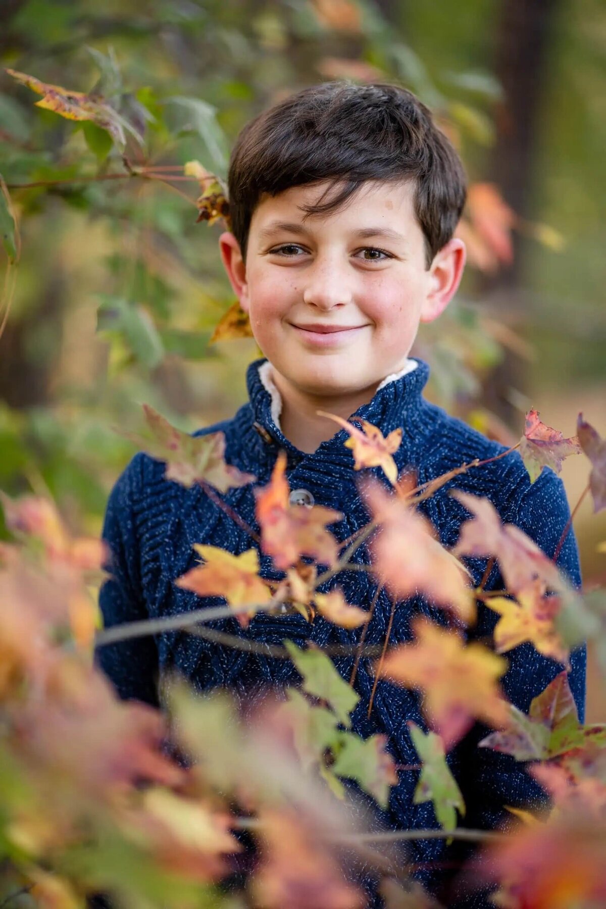 A boy smiling with fall colored trees in front of him.