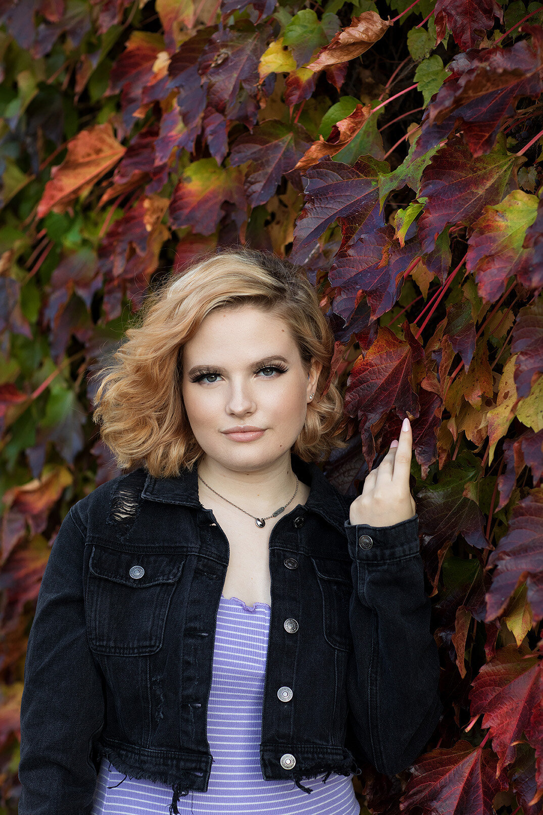 Young woman standing next to wall of colorful fall leaves wearing purple dress and black jean jacket.