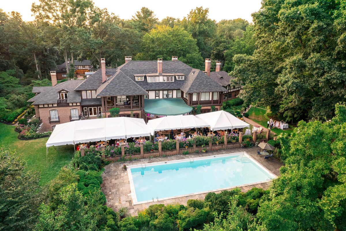 Aerial view of a large estate with a Tudor-style mansion surrounded by lush greenery. A white tent is set up in the garden next to a rectangular swimming pool. Numerous people are gathered under the tent, suggesting an event or celebration.