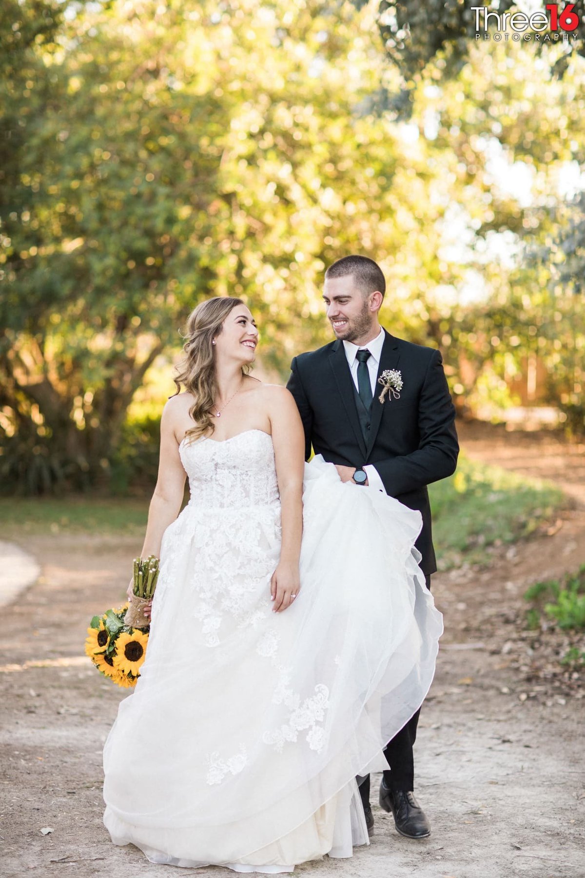 Bride looks back at her Groom as he carries her dress train while walking across a field of dirt