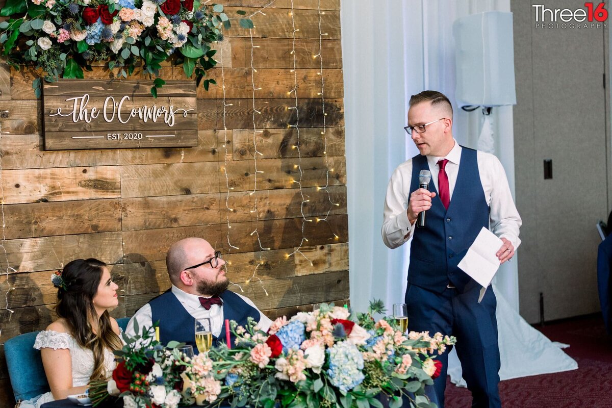 Best man delivers his toast to the newly married couple as they sit at the sweetheart table with a wood design behind them