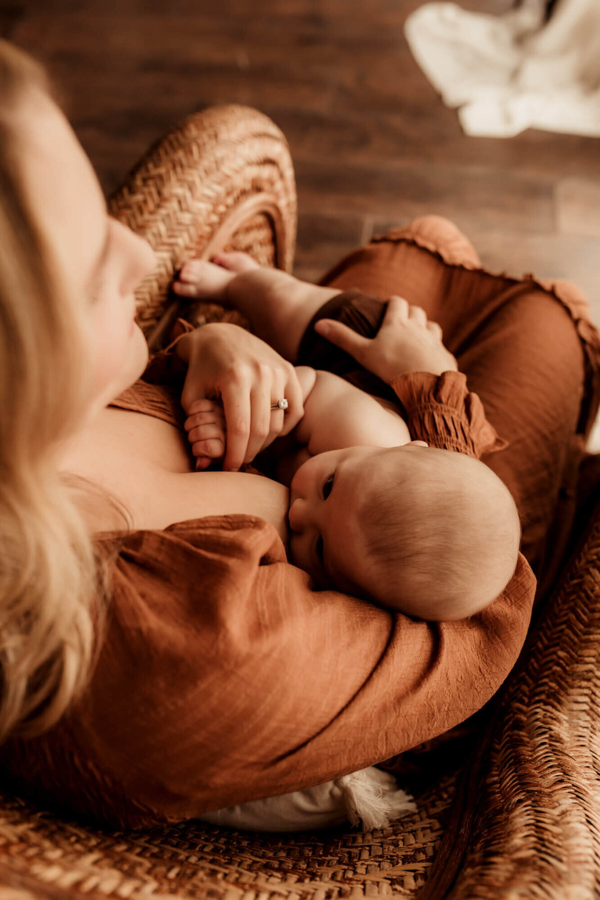 Mother nurses her son while holding his hand and sitting in a rattan chair, as she looks out the window.