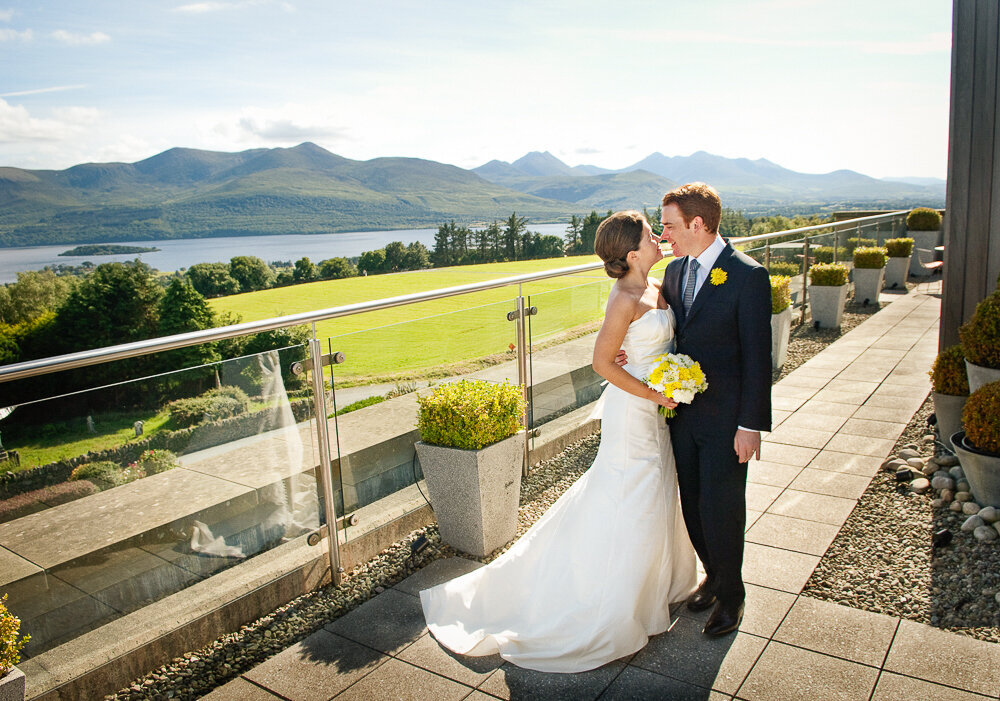 bride wearing a Greek style wedding dress, holding a yellow flower bouquet with her groom wearing a dark navy suit and yellow buttonhole, embracing on the terrace of the penthouse suite of the Aghadoe Hotel