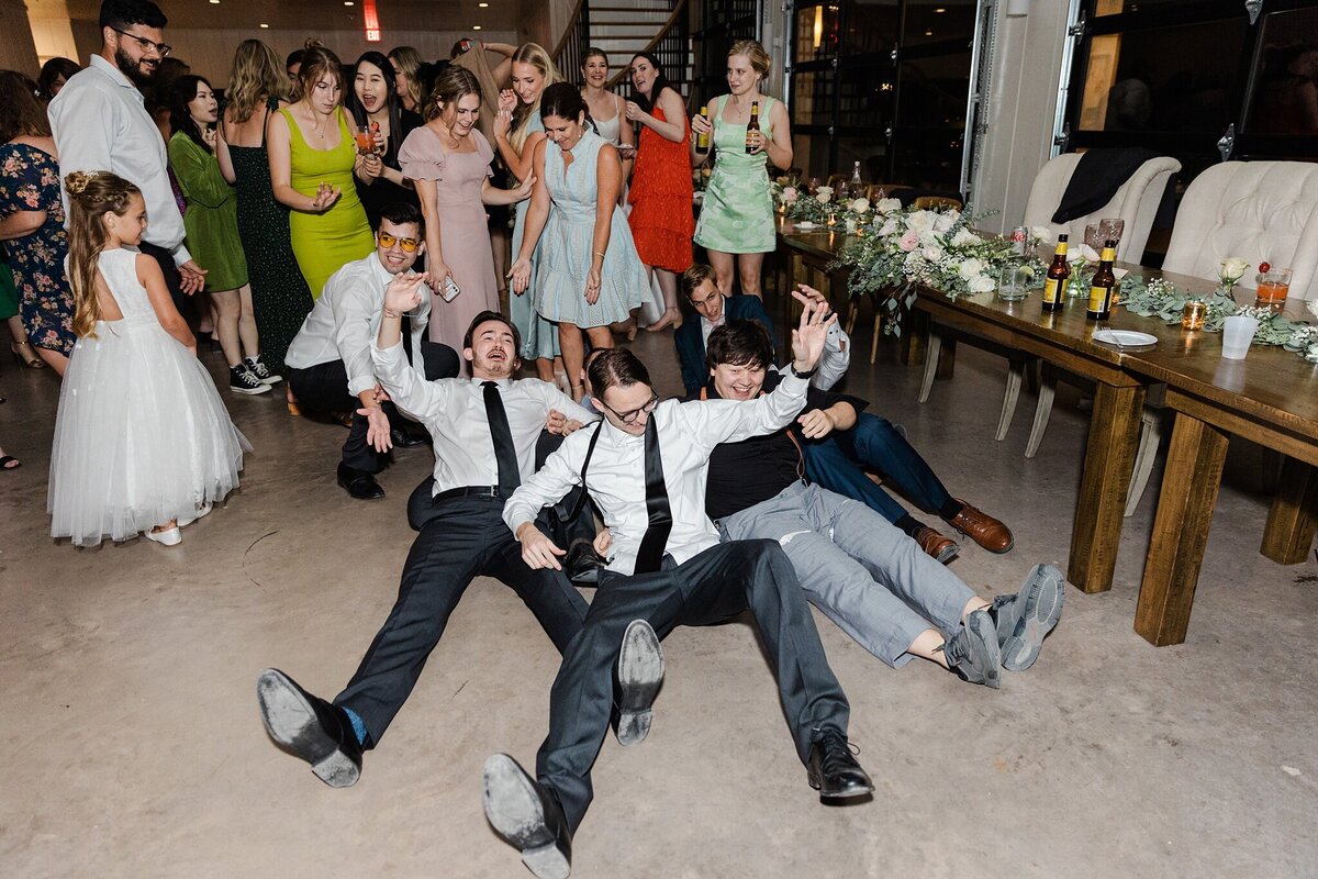 Photo of wedding guests goofing around and having fun on the floor of a wedding reception at Bella Cavalli Events in Aubrey, Texas. Five men are laughing and almost laying on the floor while many wedding guests watch joyfully behind them.