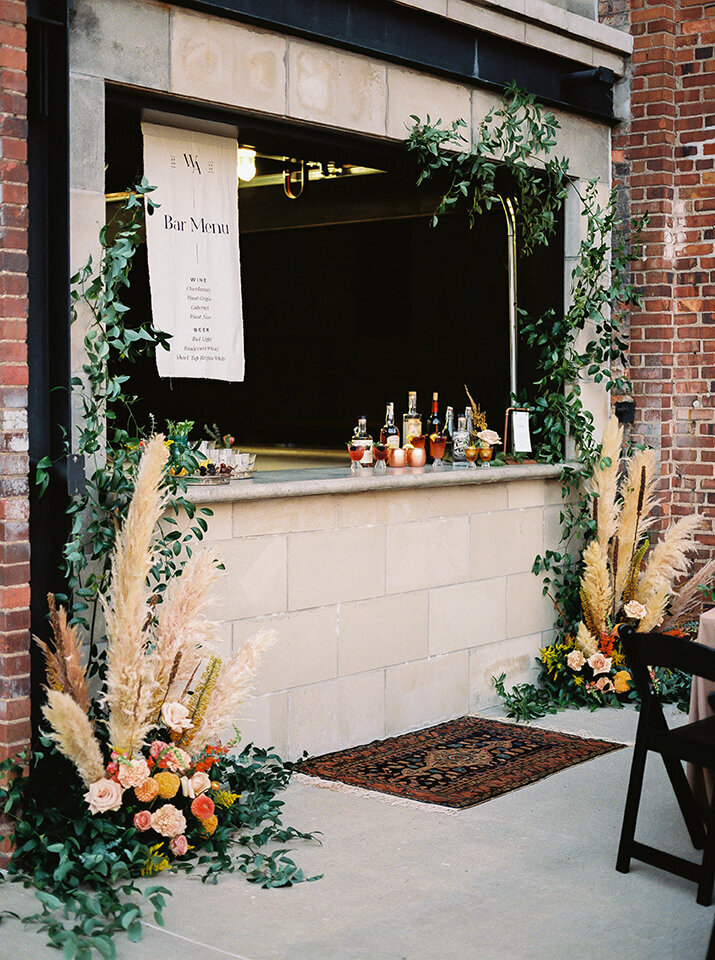 A white drink menu with black font hangs off the ceiling in the bar area outside, surrounded by flower arrangements.