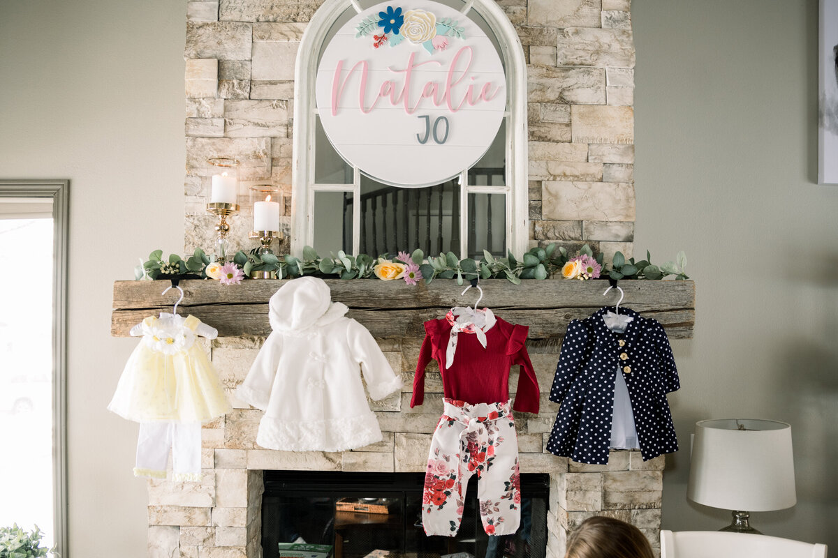 Wooden sign displaying the baby’s name hangs over a fireplace mantel adorned with greenery, candles, and baby girl outfits at a pink and yellow baby shower.