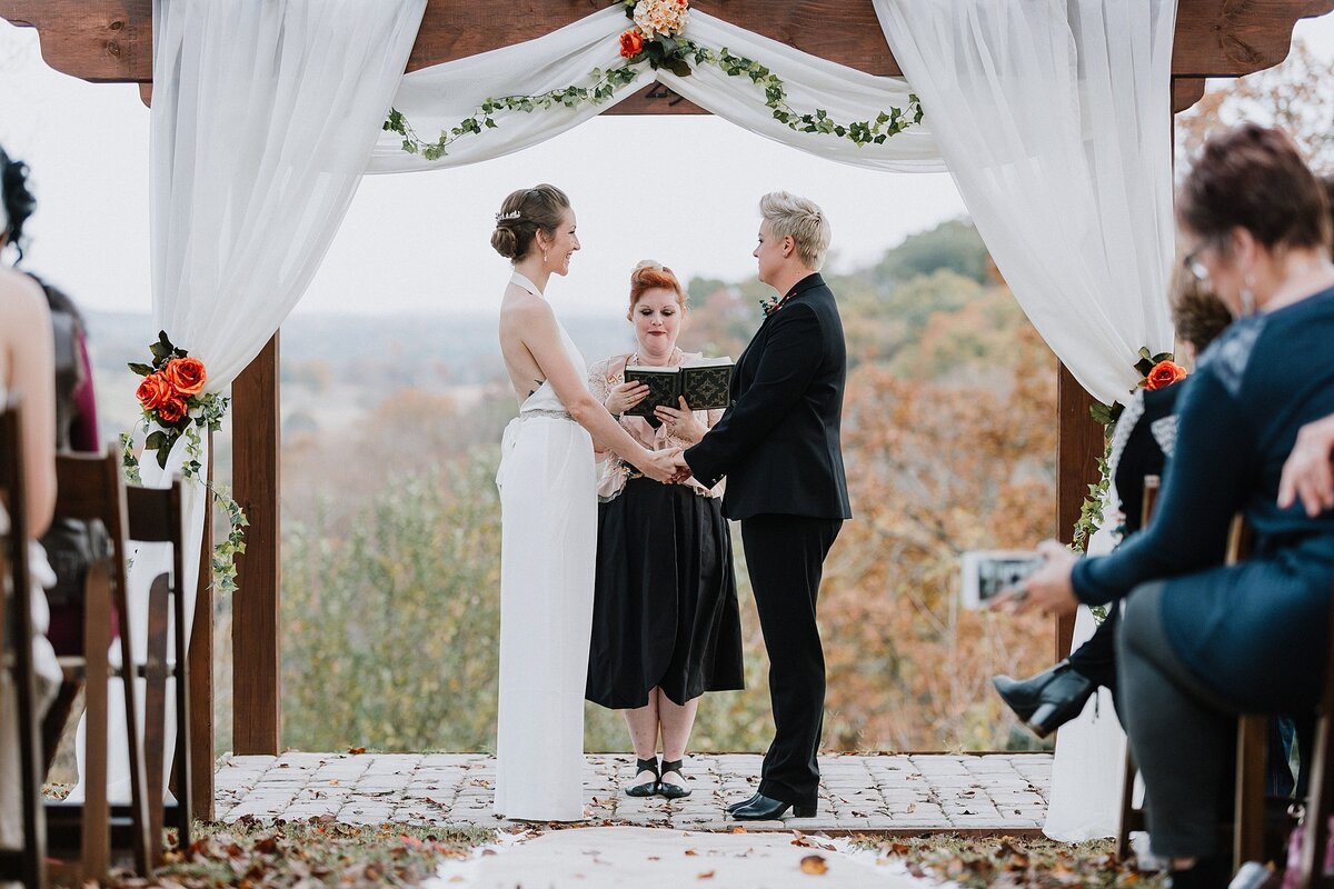 Two brides stand under a wooden arbor dressed with white drapery, ivy garland, orange roses and blush roses on a hill top in Nashville for their autumn wedding. One bride is wearing a black suit as she holds hands with  her wife who is wearing a backless white jumpsuit. The officiant is wearing a black dress and is holding a brown leather bound book.