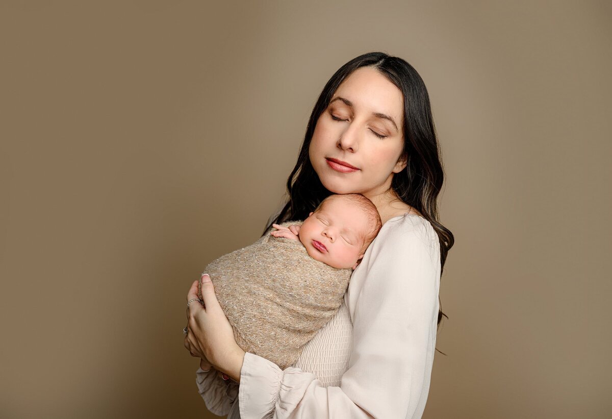 mother dressed in cream holds her newborn son wrapped in brown against her shoulder 