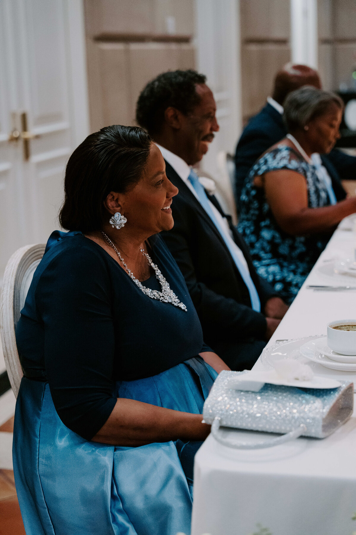 A woman in a blue dress and pearl accessories sits at an elegantly set dining table, smiling. Others in formal attire sit nearby, creating a joyful atmosphere.