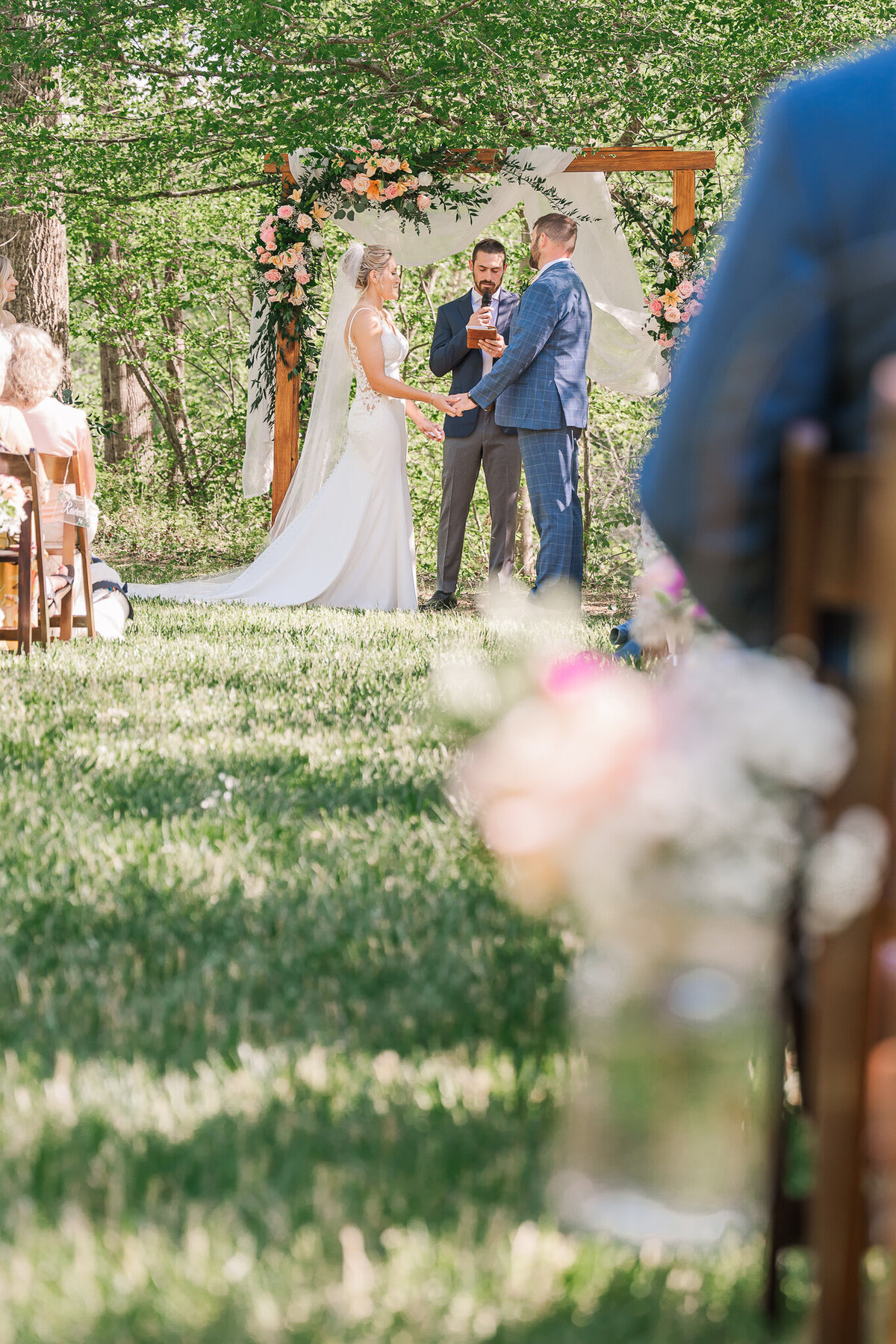 A couple exchanging vows at their family home in the Raleigh countryside under a wedding arch covered in pink flowers during the summer by JoLynn Photography, a Raleigh wedding photographer