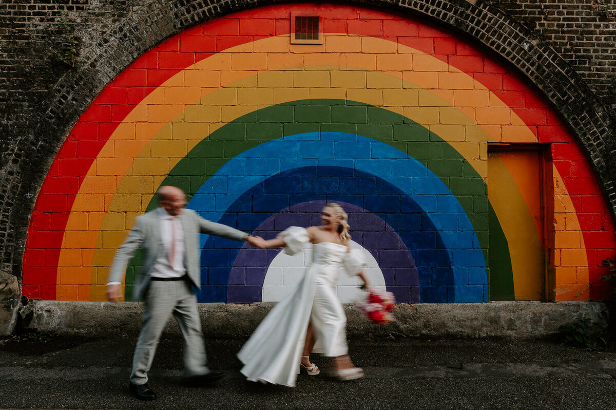 A bride pulls her groom past the rainbow mural outside 100 Barrington in Brixton, London. They have a slight blur due to a slower shutter speed. The bride is caring a pink bouquet.