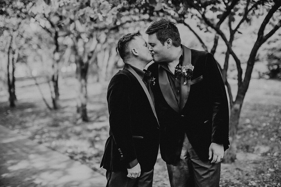 Two grooms wearing tuxedos share a kiss outdoors with trees in the background.