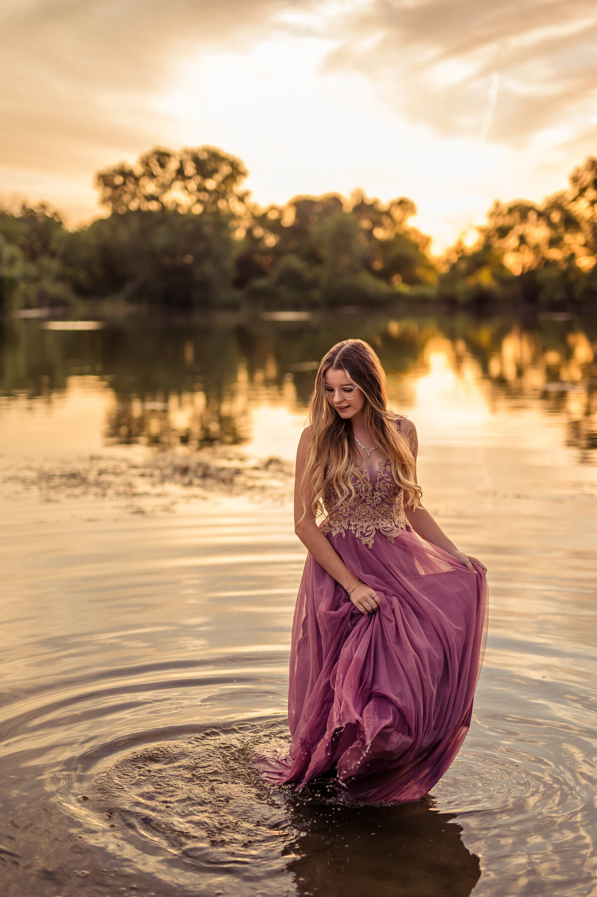 A girl from New Berlin West High School get her senior photos taken while stands in a Pewaukee lake wearing a pink and gold gown as water ripples around her.