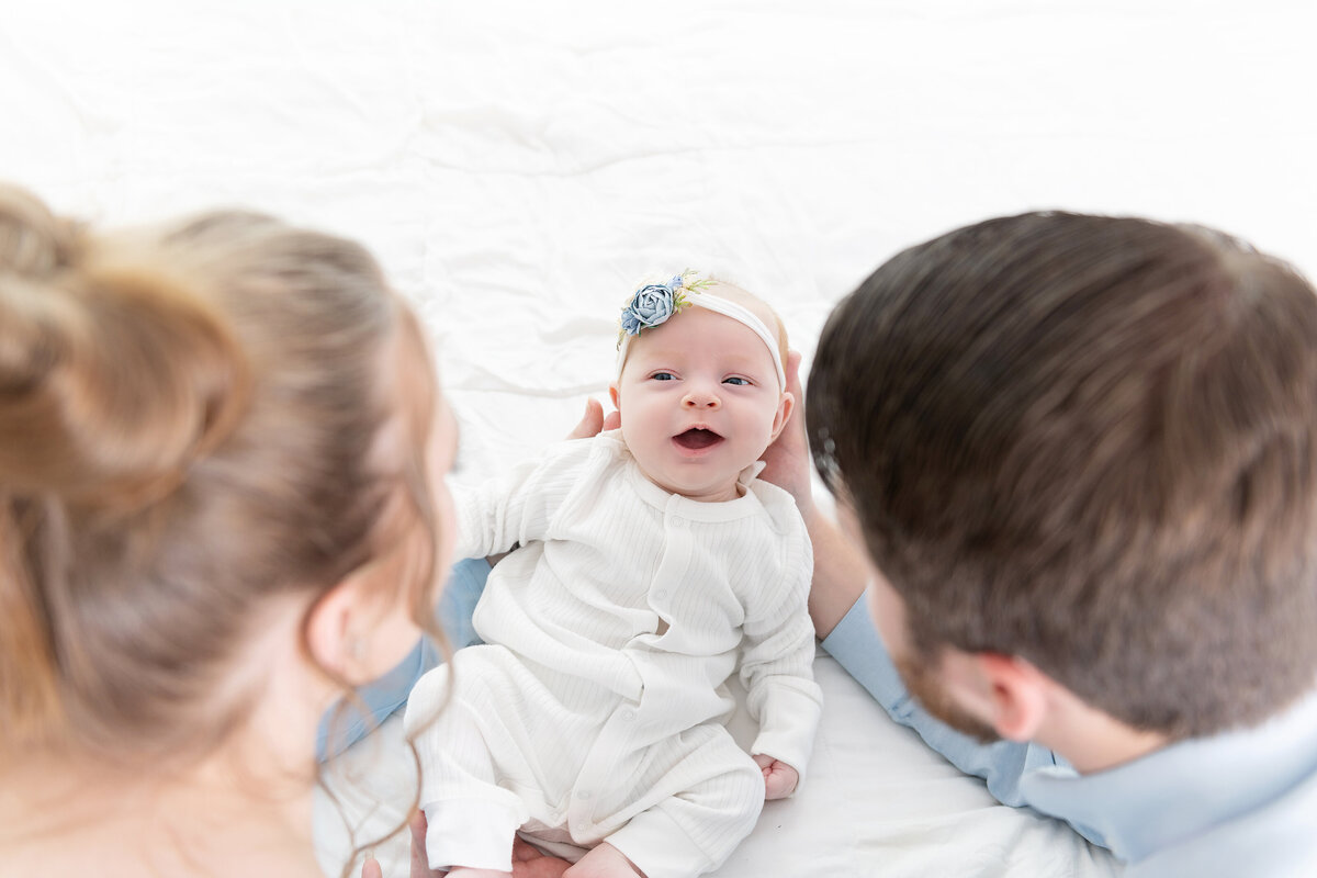 A mother and father look down at their smiling newborn baby daughter on a bed in a white onesie and blue flower headbandAtlanta newborn photographer