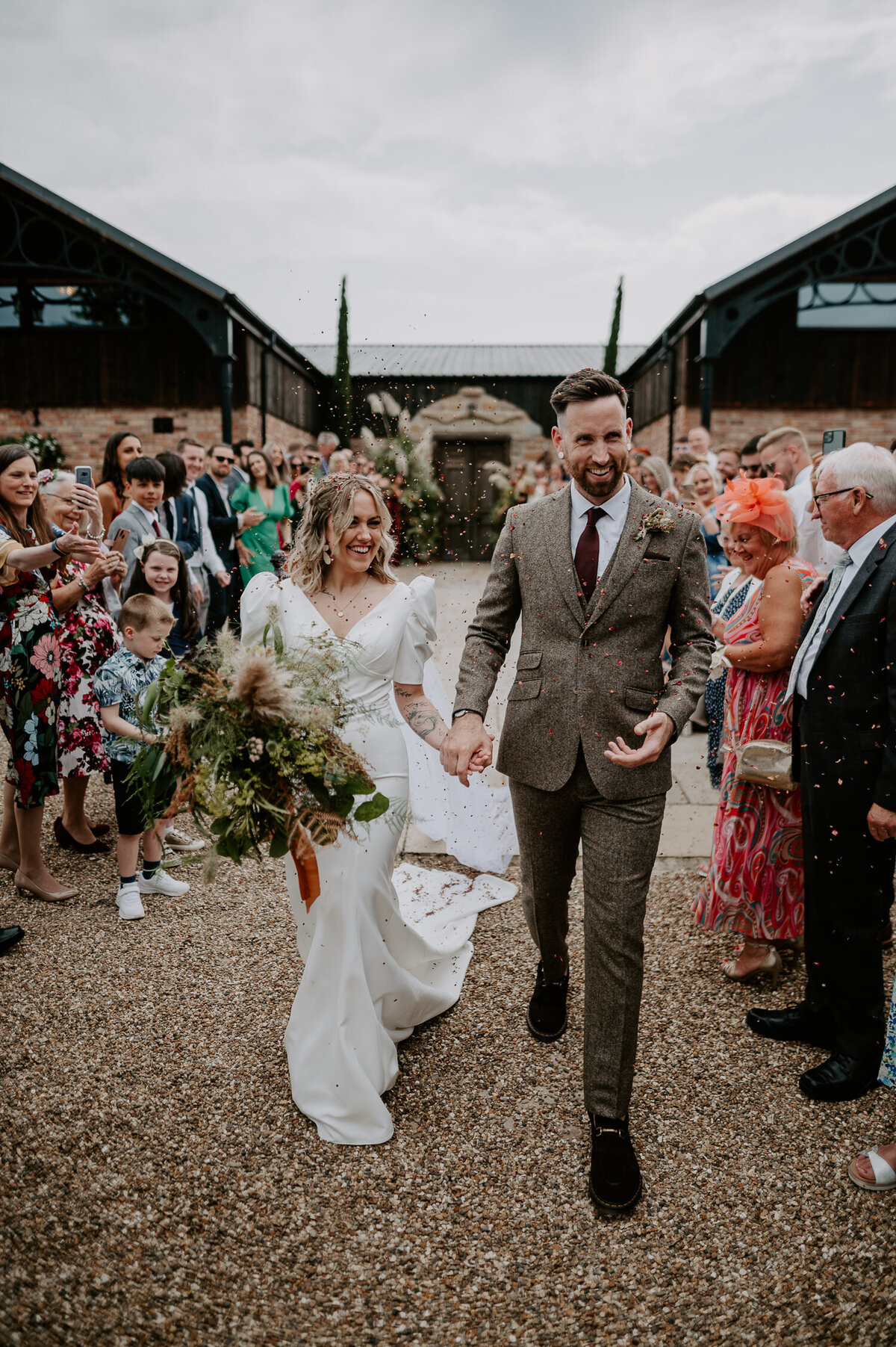 A wedding couple walk through a confetti line at The Willow Marsh Farm. The bride is wearing a puffed shoulder wedding dress and holding a large wild flower boho bouquet.