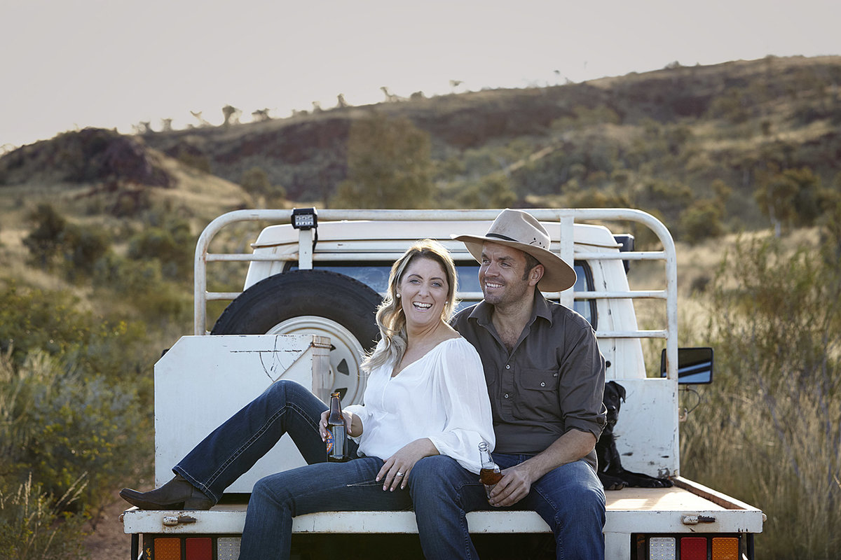 Young couple with beers in hand sitting on the back of the ute in natural bushland and with mountains in the backgroun