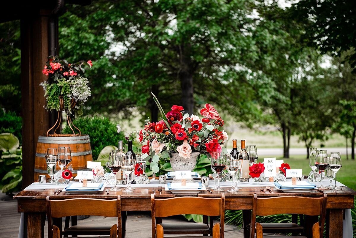 A dark wood farm table with a large blush and red floral centerpiece featuring red ranunculus, blush roses, red anthurium, red roses, red ranunculus, red anemones, blushing bride protea, blue thistle, hypericum berries, red cascading dianthus and assorted greenery at a summer wedding at Arrington Vineyards. The farm table is also set with dark wood garden chairs, white square plates and teal linen napkins. There is a large red and blush floral centerpiece on the wine barrel in the background with the vineyard vines of Arrington Vineyards in the background.