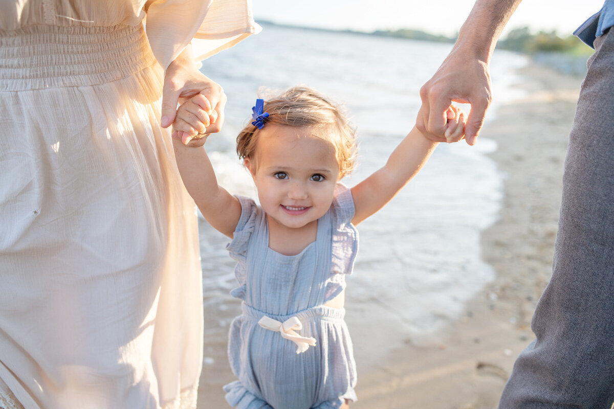 Family session of little girl holding hands with her parents at the beach