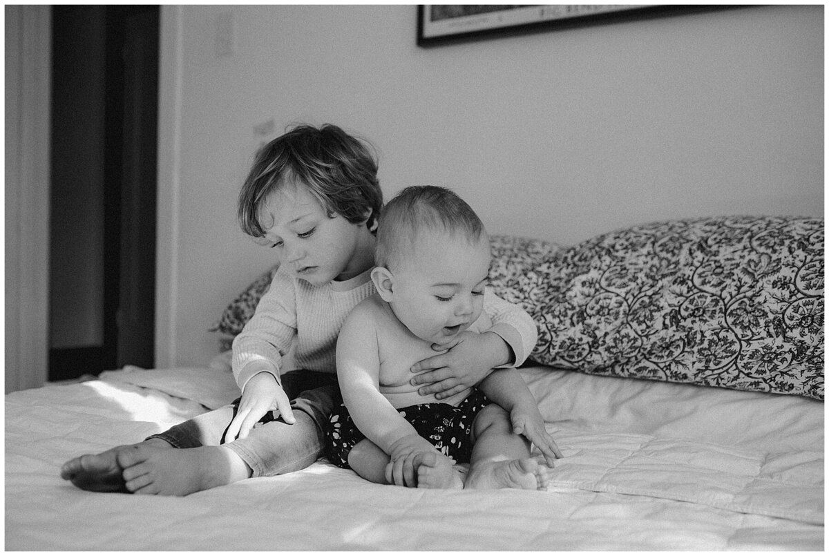 Toddler sitting on bed looking at a toy holding baby brother at sweet home family session in Austin by Amber Vickey Photography