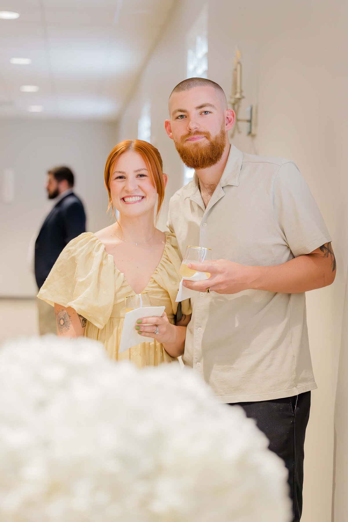 A couple stands in a well-lit room, captured by an event photographer in Fayetteville, NC, holding drinks and napkins with warm smiles. Dressed in light-colored outfits, they pose as a suited figure appears faintly in the background and white flowers partially obscure the foreground.