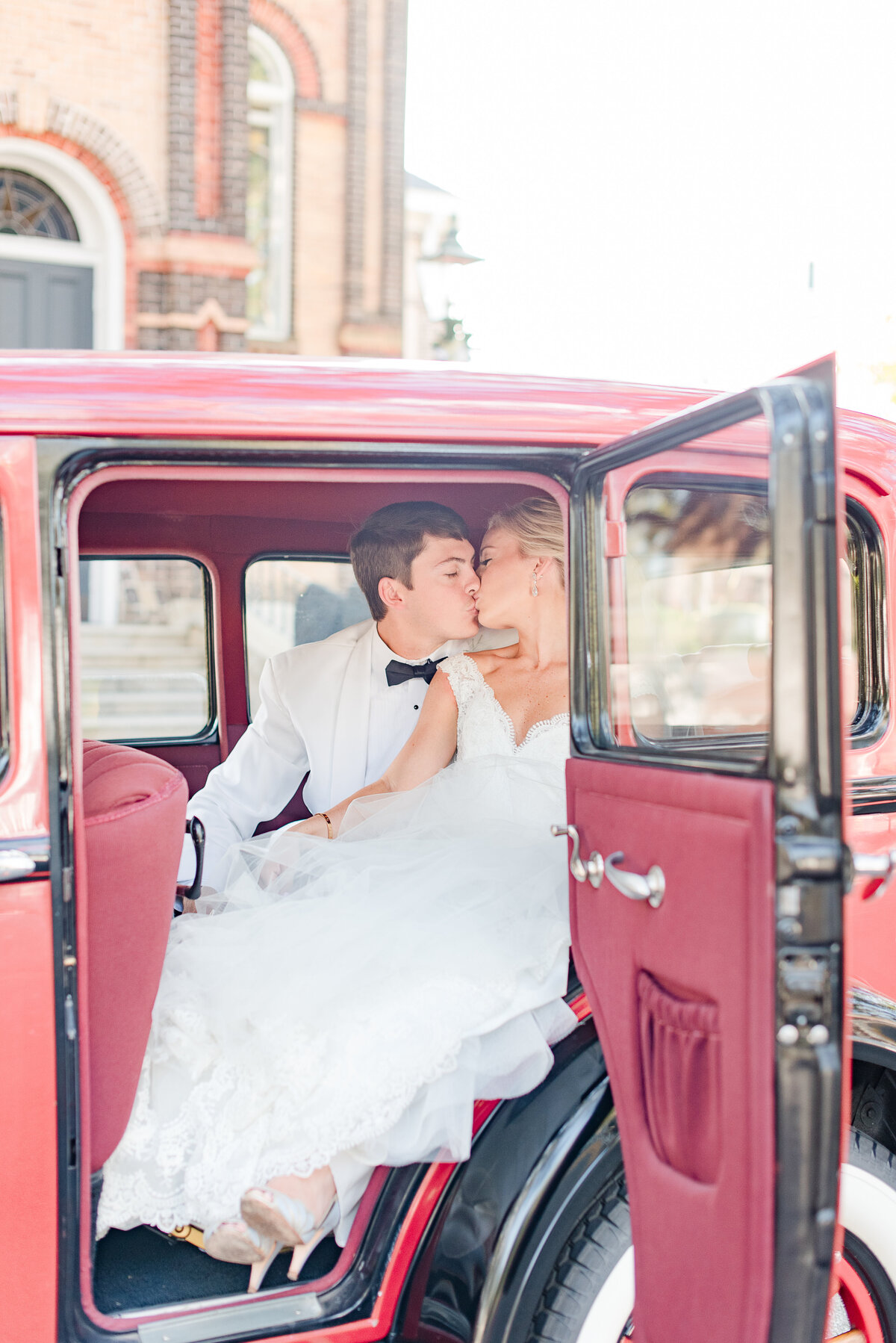 couple kissing inside red classic car on wedding day white tuxedo