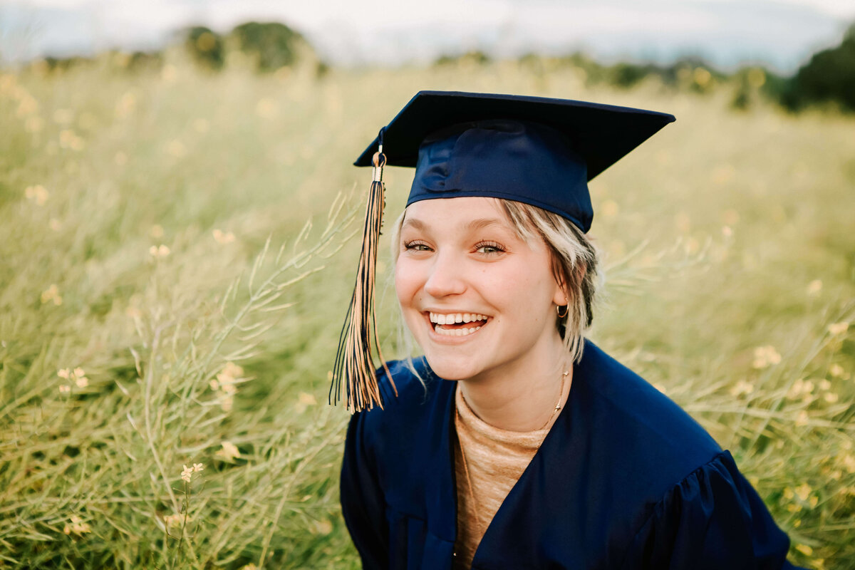 A West Albany High School senior girl looks at the camera smiling, posing for senior portraits with Jen White Photography. She's in a dark blue cap and gown with gold sweater under and gold and blue tassell