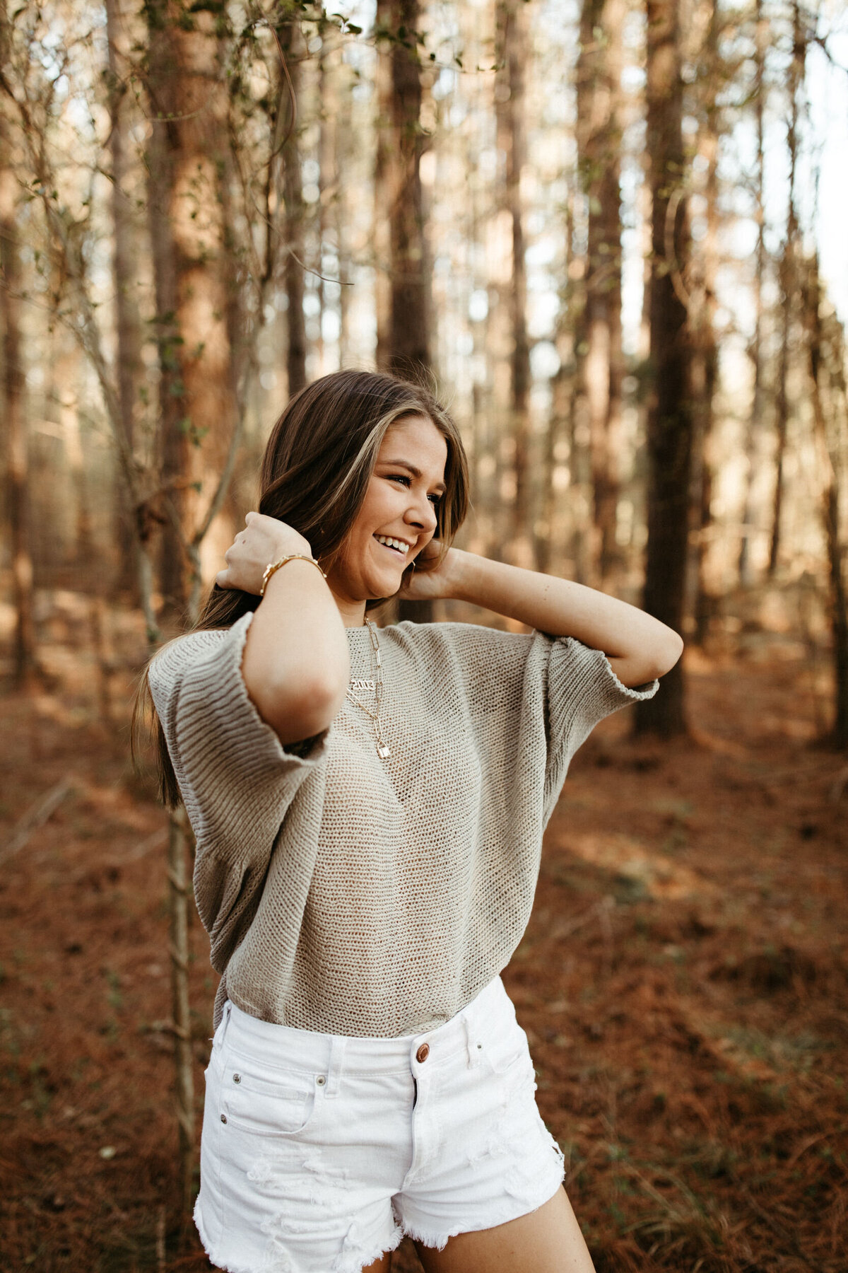 Senior girl in white shorts and grey loose shirt smiling in the woods